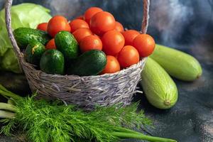 Fresh vegetables for salad in a basket. Tomatoes and cucumbers with zucchini and cabbage with dill. Spring harvest, benefits and vitamins. On a dark background. photo