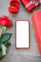 White blank screen of phone with gift boxes, rose and ring on wooden background. photo