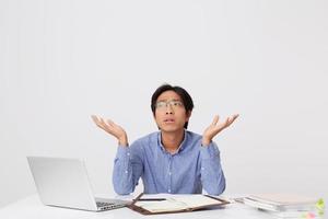 Confused thoughtful asian young business man in glasses sitting at the desk with laptop and notebook and holding copyspace on both palms over white background photo