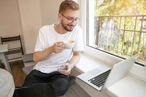 toma interior de un joven barbudo encantador, sentado junto a la ventana con el teléfono en la mano y una computadora portátil en el alféizar de la ventana, tomando café y mirando la pantalla con una sonrisa foto