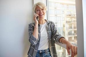 Happy young woman with blond short hair in casual clothes talking on phone with friend, laughing at joke, leaning on window and looking aside photo