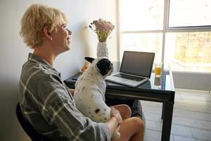 mujer joven independiente que trabaja lejos de casa usando una computadora portátil, sentada en la mesa al lado de la ventana y sosteniendo a su perro, estando de buen humor foto