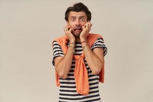 Shocked scared young man with stubble in striped t shirt and sweater on shoulders feels terrified and looks frightened over white background photo