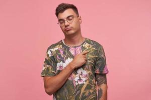 Charming young man with trendy haircut standing over pink background with eyes closed, keeping hand on his chest, being calm and balanced photo