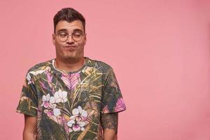 Funny pretty young male in flowered t-shirt and glasses looking aside indifferently, folding lips and making apathetic face, standing over pink background photo