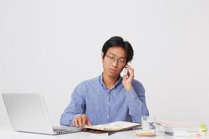 Atractivo y exitoso hombre de negocios asiático con anteojos y camisa azul que trabaja con una laptop y un portátil hablando por teléfono celular en la mesa sobre fondo blanco. foto