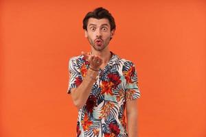 Happy charming young man with bristle in colorful shirt standing and sending a kiss to camera over orange background photo