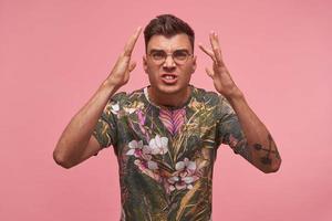 Indoor shot of angry short haired man in flowered t-shirt and glasses looking to camera crossly, raising his palms with perplexity, isolated over pink background photo
