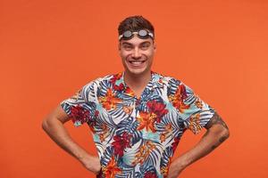 Happy attractive young swimmer with goggles on his forehead standing over orange background, looking at camera joyfully with hands on hips photo
