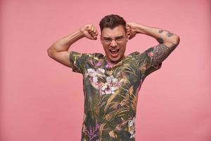 Strong handsome young guy with tattoos looking smugly to camera, raising hands up and demonstrating his muscles, posing over pink background photo