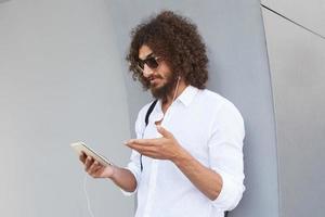 Atractivo joven barbudo de cabello oscuro y rizado apoyado en una pared exterior gris durante una videoconferencia con una tableta, con gafas de sol y ropa informal foto