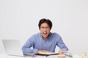 Angry furious asian young business man in glasses working at the table with laptop screaming and looking at camera over white background photo