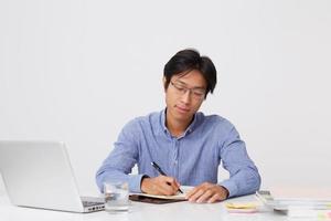 serio y exitoso joven hombre de negocios asiático con anteojos trabajando con una laptop y escribiendo en un cuaderno en la mesa sobre fondo blanco