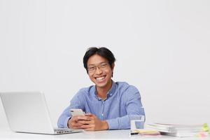 Happy smiling asian young business man in glasses using cell phone laughing and working at the table with laptop over white background photo