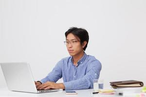 un joven hombre de negocios asiático serio y centrado con gafas sentado en la mesa escribiendo y trabajando con un portátil aislado sobre fondo blanco foto