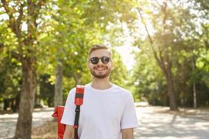 Portrait of joyful handsome guy with stubble, wearing sunglasses and casual clothes, standing over green park on nice sunny day, looking happy and pleased photo