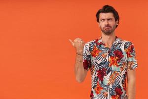 Sad disappointed young man with stubble in hawaiian shirt standing and pointing to the side at empty space by thumb finger over orange background photo