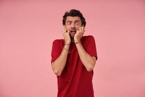 Hysterical crazy young man with stubble in red t shirt feels terrified and looks panicked over pink background photo