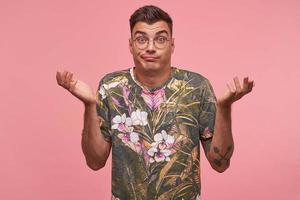 Bewildered attractive short haired guy with glasses raising palms up with complicated face expression, contracting forehead, posing over pink background photo