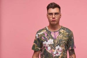 Offended pretty short haired young male in glasses looking aside with resentment, being upset about something, making pained face, posing over pink background photo