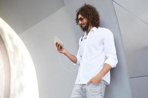 Young curly dark haired man with beard leaning on grey wall, posing over city background, having video calling and talking to somebody using headphones, smiling to tablet camera photo