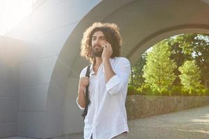 Pretty young curly man with beard walking through city green park on warm sunny day, giving call with his smart phone, wearing white shirt and black backpack photo