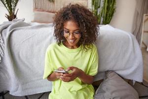 Happy young dark skinned curly female holding smart phone in hands, checking her social networks, looking at screen and smiling, posing over home interior