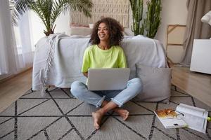 Cheerful young dark skinned woman sitting on floor in casual clothes, studying with textbooks and modern laptop, smiling joyfully and being in nice mood