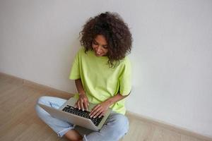 Indoor photo of happy young curly woman with dark skin chatting with friends on laptop, sitting with crossed legs, wearing jeans and yellow t-shirt