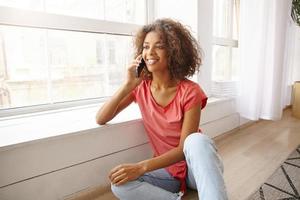 Indoor portrait of lovely curly female with dark skin having pleasant conversation on mobile phone, looking ahead and smiling joyfully, sitting near window photo