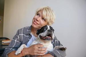 Close-up portrait of cheerful black and white dog, sitting in a hug, posing over blond beautiful young female in casual clothes, looking dreamily aside photo