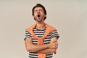 Tired exhausted young man with bristle in striped t shirt and sweater on shoulders keeps arms crossed and yawning over white background photo