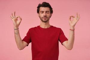 Relaxed concentrated young man with stubble and closed eyes in red t shirt showed drama gesture and practicing yoga and meditation over pink background photo