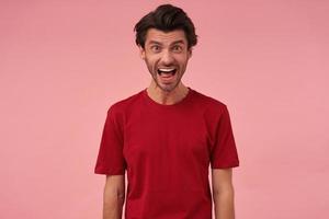 Portrait of crazy angry young man with bristle and opened mouth in red t shirt looks mad and screaming over pink background photo