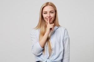 Horizontal shot of beautiful blonde long haired female keeping finger on smiling lips, demonstrating hush sign, asking to keep silence, standing over white background photo