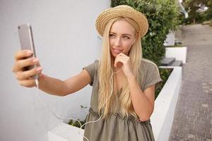 Young attractive blonde woman making selfie with her smart phone, looking to phone camera gently and touching her chin with hand, wearing casual linen dress and straw hat photo