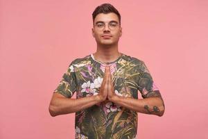 Portrait of calm handsome male in casual t-shirt standing over pink background with palms together at his chest, making namaste gesture, going to do yoga photo