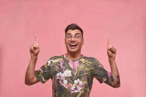 Studio photo of charming young short haired male in flowered t-shirt looking to camera joyfully, pointing with fingers upwards, isolated over pink background
