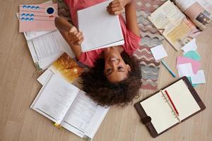 Top view of attractive young curly female with dark skin lying on carpet while reading notes, preparing for exams with a lot of books, wearing pink t-shirt photo