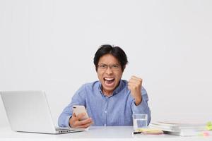 Angry aggressive asian young business man in glasses working with laptop and mobile phone shouting and showing fist gesture over white background photo