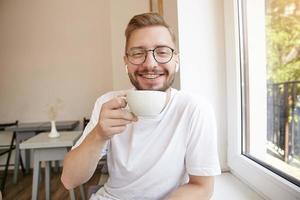 Indoor close-up of cute happy bearded guy, holding a cup in his hand and looking to camera with wide smile, wearing white t-shirt and glasses photo