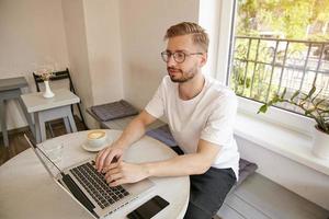 joven trabajador independiente barbudo con camiseta blanca y anteojos, sentado en la mesa en la cafetería y usando una computadora portátil, revisando correos electrónicos y luciendo concentrado foto