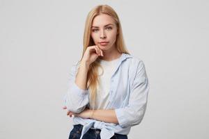 Severe attractive young woman with long blond hair wearing casual clothes standing over white background, looking serious to camera and keeping hand under chin photo