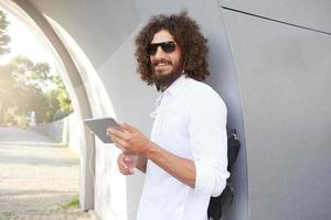Young attractive curly man with beard smiling sweetly, listening to music with earphones on his tablet, posing over outdoor background on sunny warm day photo