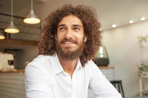 Happy handsome bearded guy with curls looking aside and smiling broadly, being in nice mood, meeting friends during lunch break, posing over coffee house photo