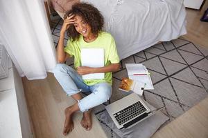 Good looking young curly woman with brown curly hair sitting on carpet with geometric print, making break with studying and looking dreamily out window photo