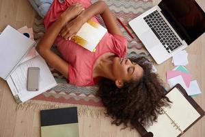 Beautiful young curly lady with dark skin lying on floor between books, notebooks and laptop, posing over colored carpet with closed eyes and pleasant smile photo