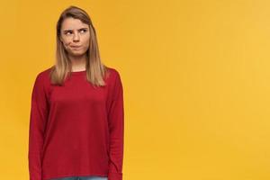 Thoughtful girl with blond hair. Wearing red sweater. Lips pursed like doubting on some idea. Free copy space for your text. Looking to the upper right corner isolated over yellow background photo