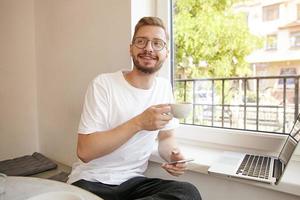 Portrait of cute young guy with beard and glasses, working remotely with laptop, holding smart phone and coffee in his hands, being positive and happy photo