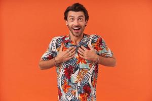 Marveled excited young man with stubble and opened mouth in hawaiian shirt keeps both hands on chest and looks surprised over orange background photo